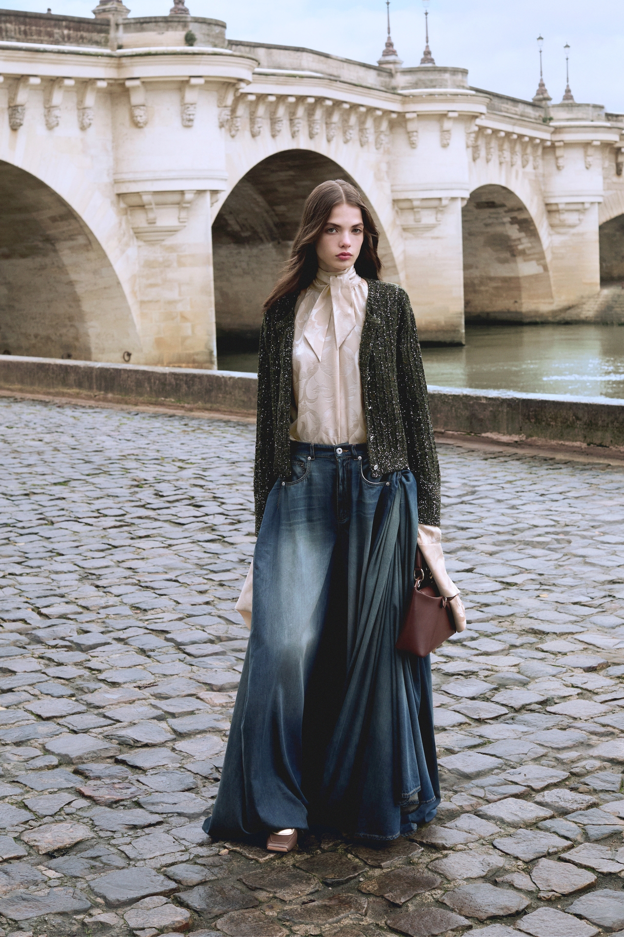 A person wearing a long denim skirt, white blouse, and dark jacket stands on cobblestone near a stone bridge over a river.