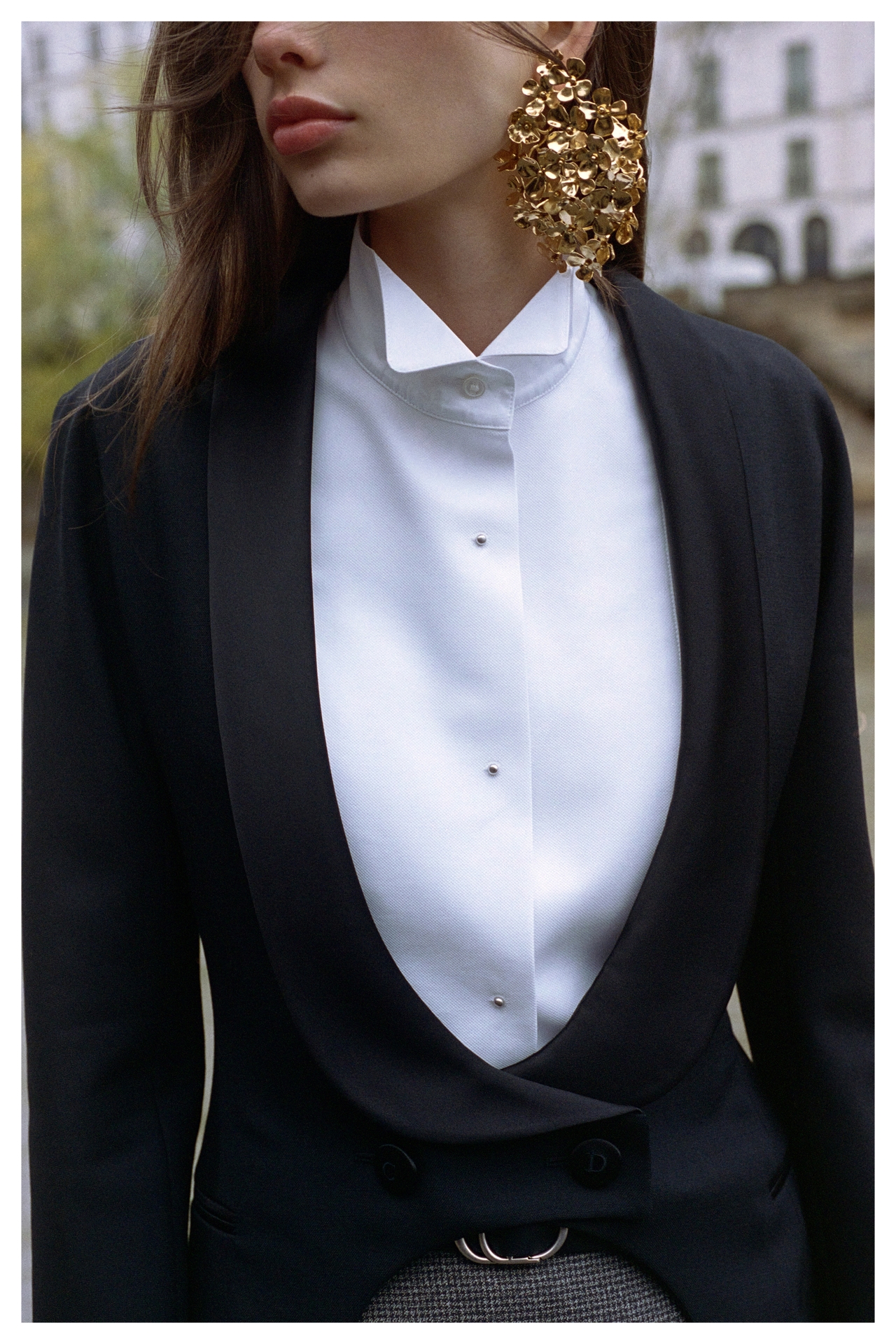 Woman wearing a black tuxedo jacket, white shirt, and large gold floral earrings, standing outdoors with blurred buildings in the background.