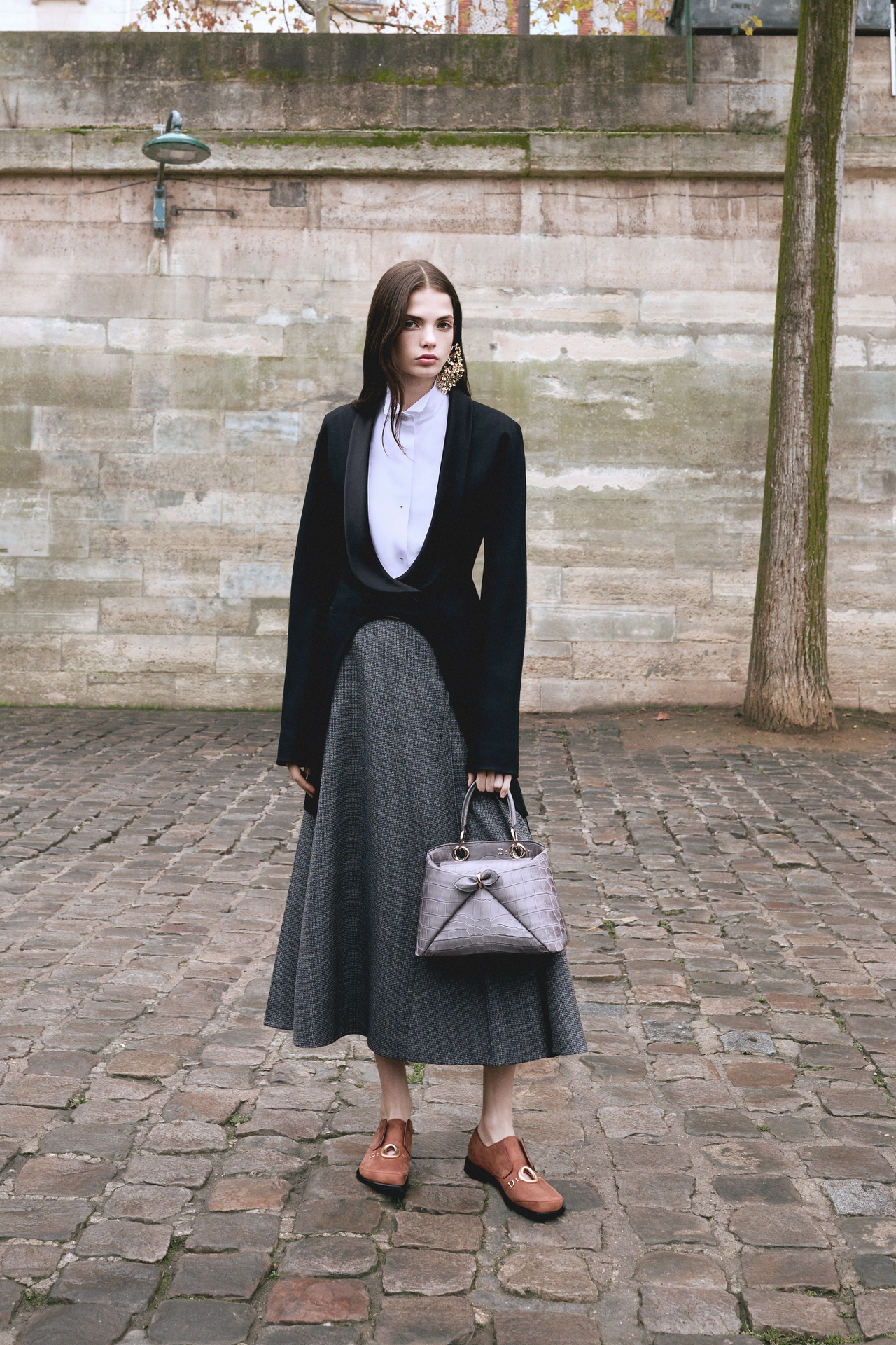 Fashionable person in a black jacket, white blouse, gray skirt, holding a textured bag, standing on cobblestone pavement against a stone wall.
