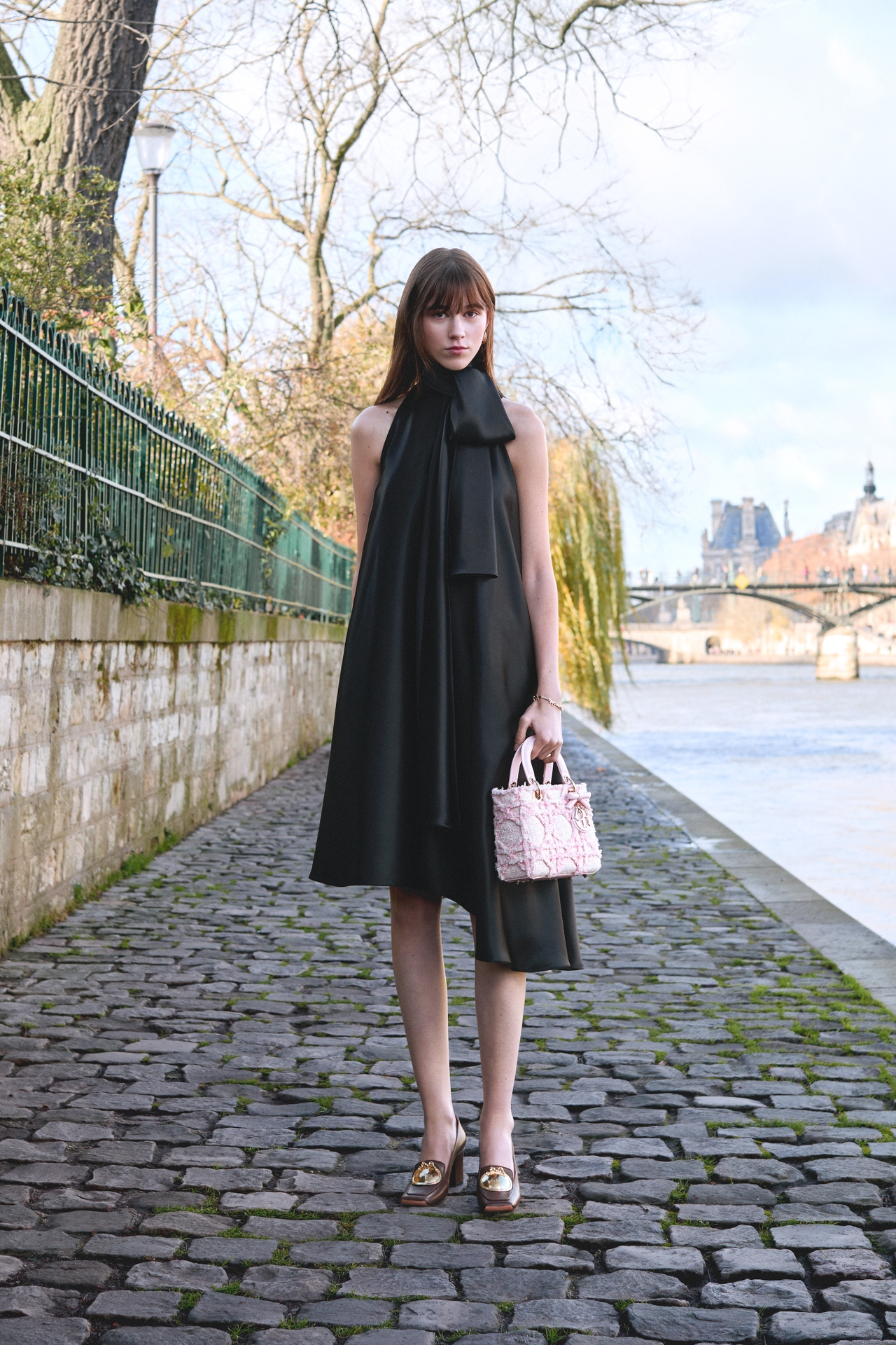Woman in a black halter dress stands on a cobblestone path by a river, holding a patterned handbag; trees and buildings in the background.