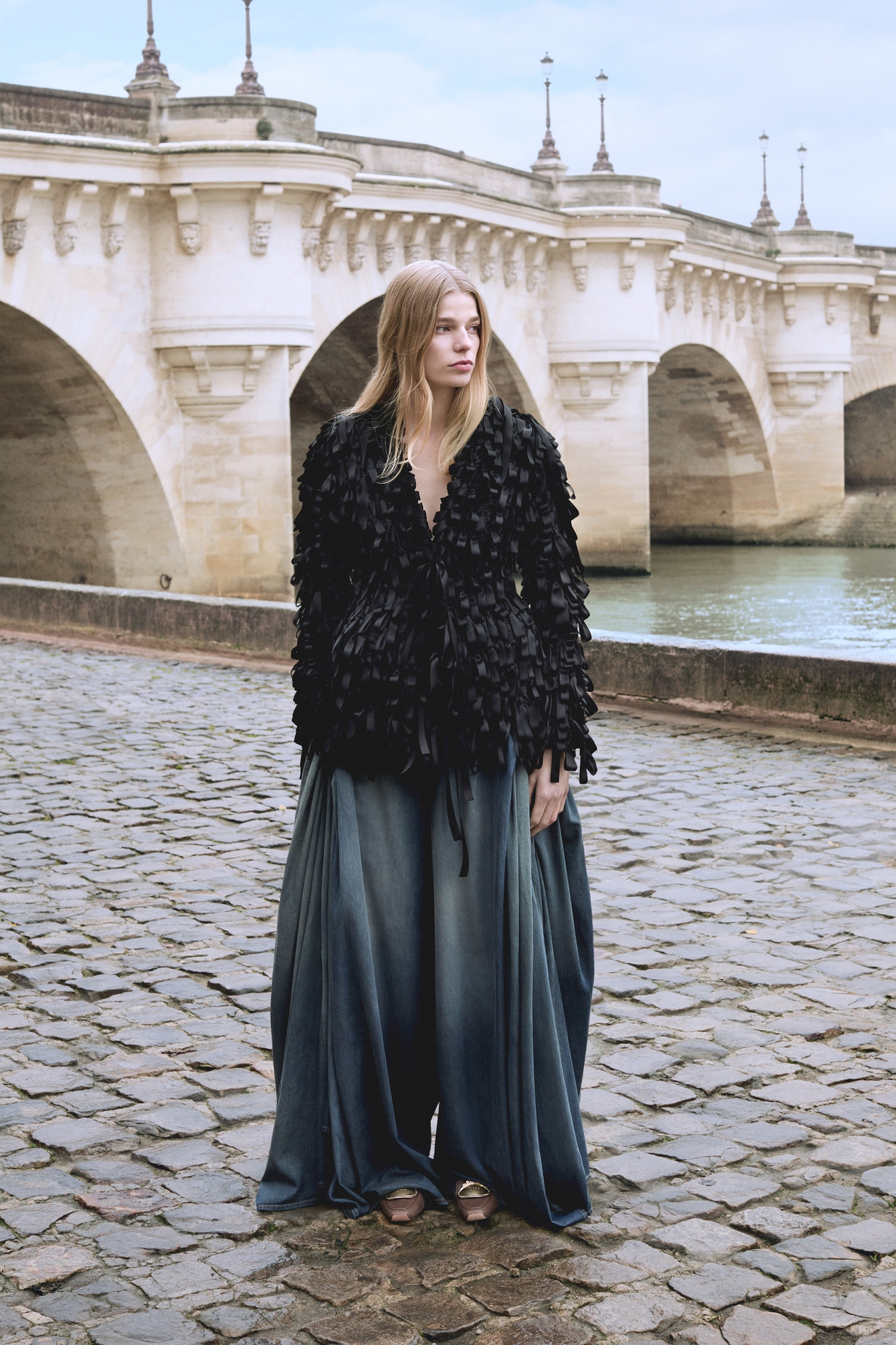 Woman in a textured black jacket and flowing pants stands on a cobblestone path near a stone bridge over a river.