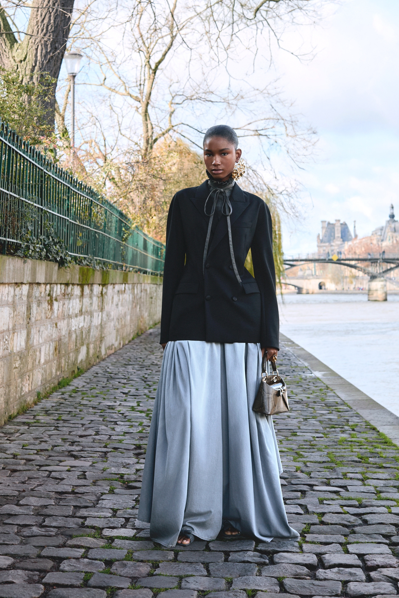 Person in a black jacket and flowing silver skirt stands on a cobblestone path by a river, holding a handbag. Trees and a bridge in the background.