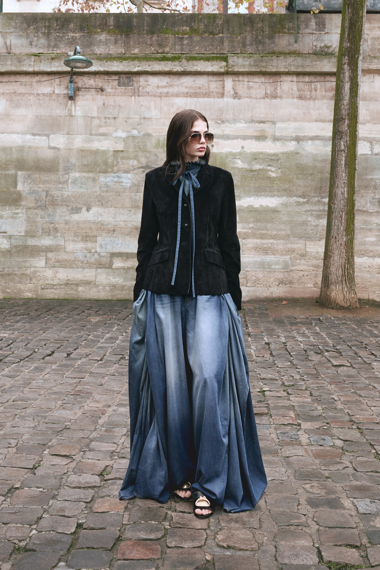 Woman in sunglasses wears a black jacket and flowing blue pants, standing on a cobblestone path against a stone wall background.