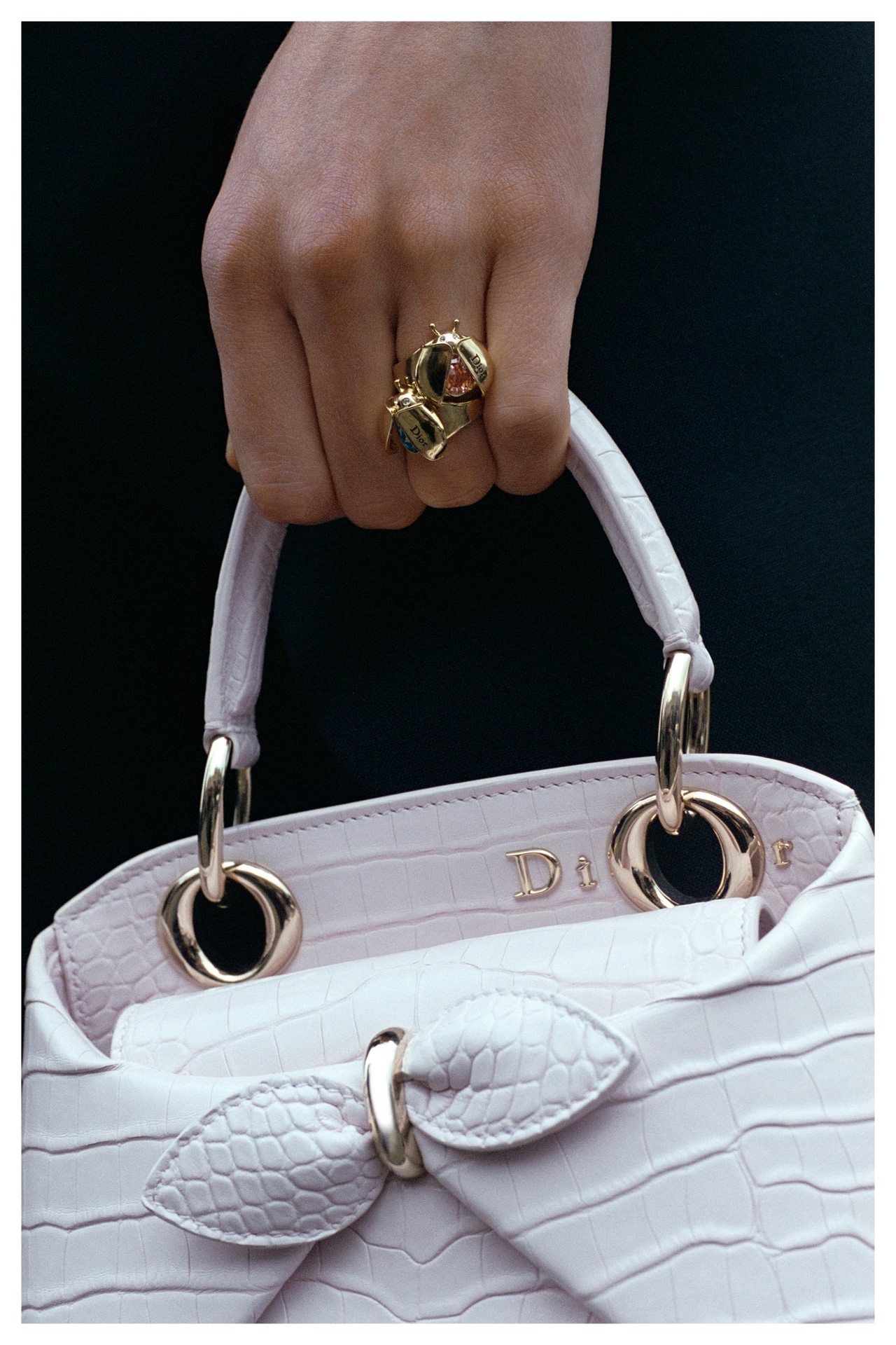 Hand holding a white textured handbag with gold-rimmed handles, wearing a chunky gold ring adorned with colorful gems.