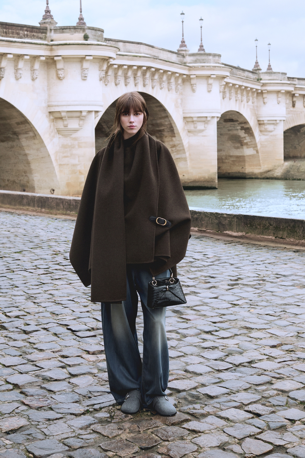 Person standing on cobblestone by a river, wearing a brown cloak and holding a black bag, with a stone bridge in the background.