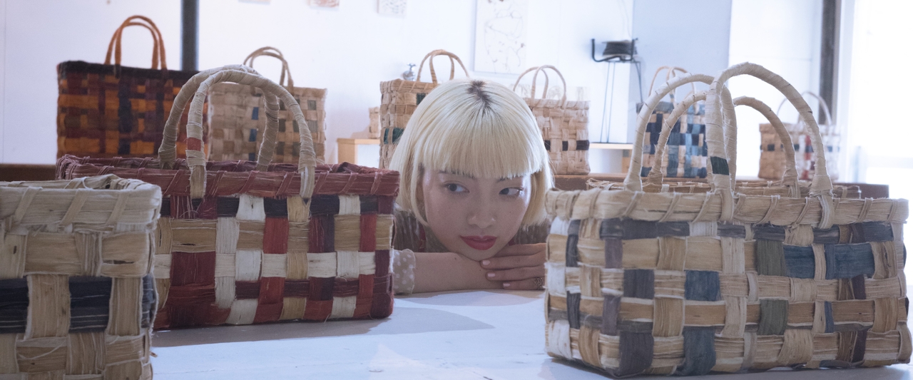 Person with blonde hair looking at woven baskets on a table, with various colors and patterns, in a well-lit room.