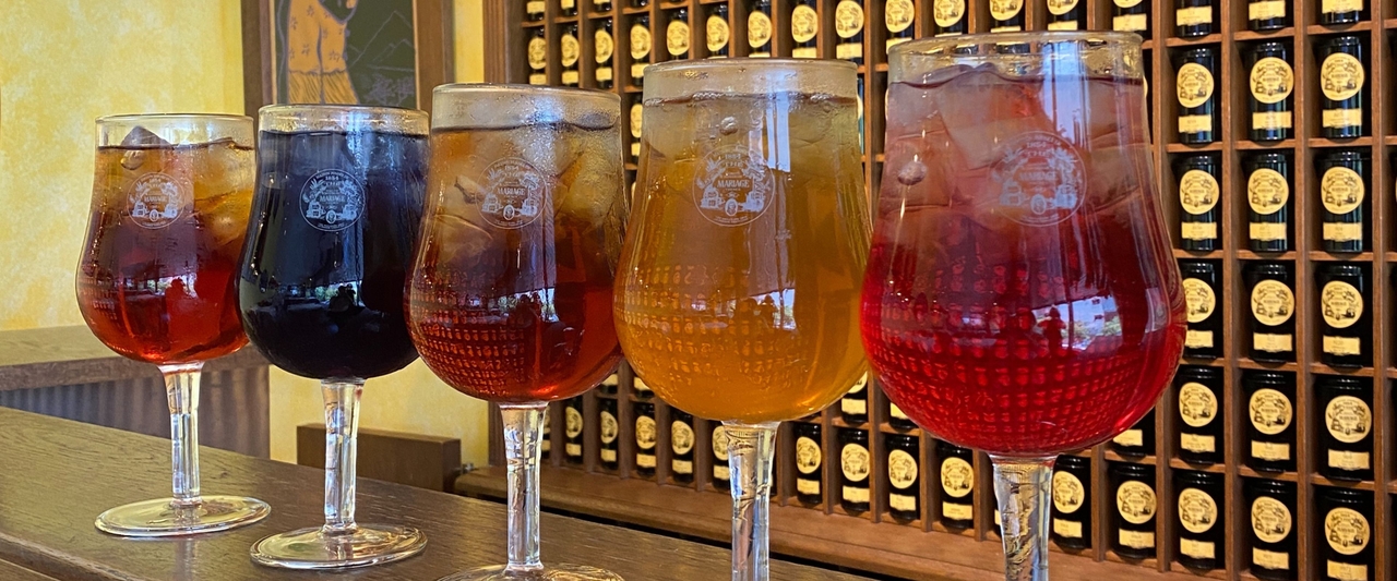 木製のカウンターの上に色とりどりのアイスティーが5杯並び、背景にはワインボトルが並んだ壁が広がっている。