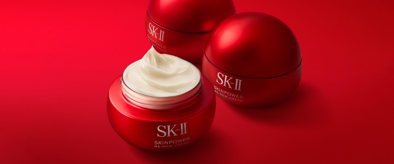 Three red SK-II skincare jars on a red background, one jar open showing white cream inside.