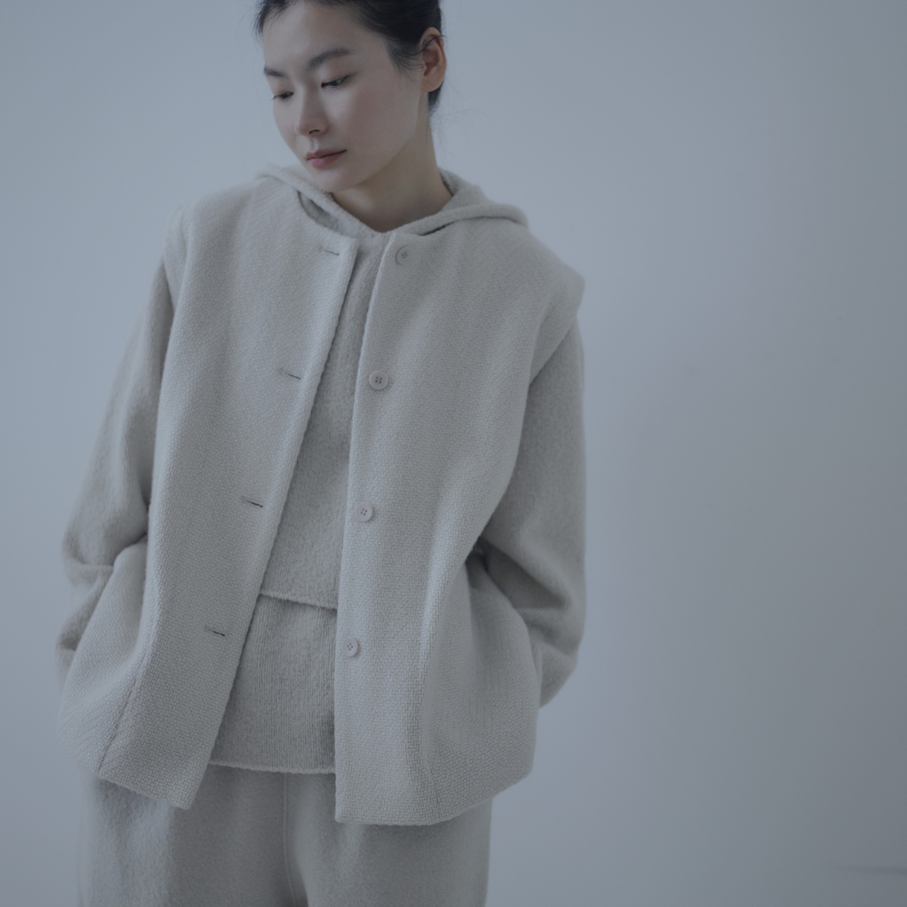 ライトベージュのフード付きジャケットと同色のパンツを着用した人物が、シンプルな背景の前でポケットに手を入れながら立っている。