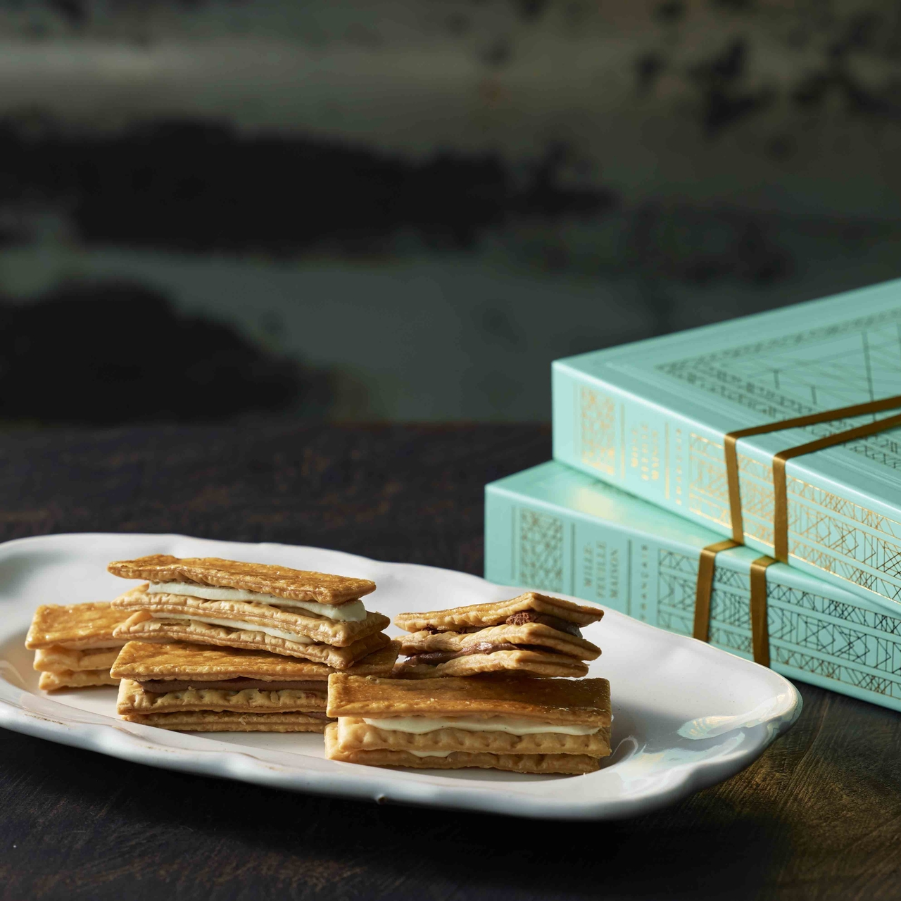A plate of layered pastry snacks on a table, with elegant light blue boxes in the background.