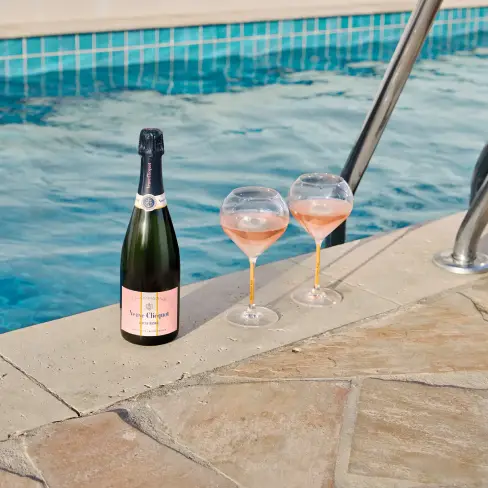 Bottle of rosé champagne, Veuve Clicquot Rich on Ice Rosé&nbsp;and two filled glasses on a poolside ledge, with clear blue water in the pool and a metal ladder in the background.