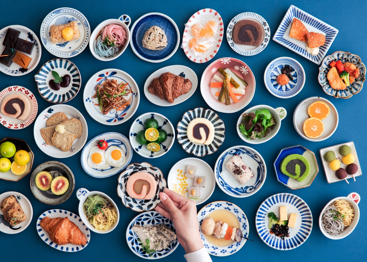 An array of colorful Japanese dishes on various plates, with a hand reaching to pick up a dessert.