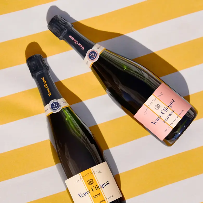 Two bottles of Veuve Clicquot champagne, Veuve Clicquot Rich on Ice and Rich on Ice Rosé, one with a yellow label and the other with a pink label, on a yellow and white striped background.
