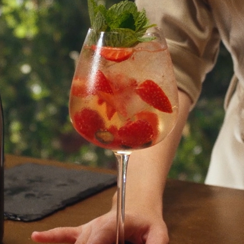 A glass of iced champagne cocktail, Golden Hour with sliced strawberries and mint leaves, held by a person in a light shirt.