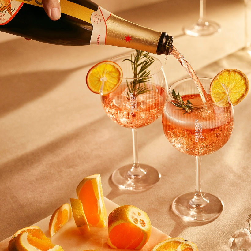 Champagne, a Chandon Garden Spritz being poured into two glasses garnished with orange slices and rosemary, with sliced oranges on a cutting board nearby.